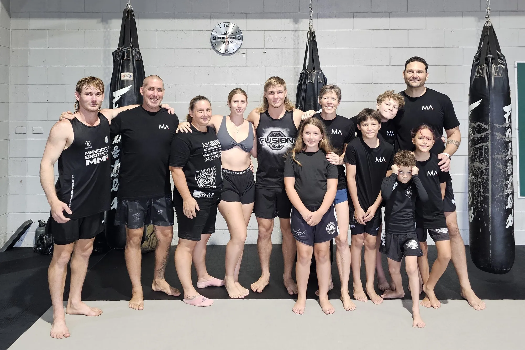The Maunder and Mayocchi families standing together on the mats at The M A Connection Innisfail, in front of the heavy bags